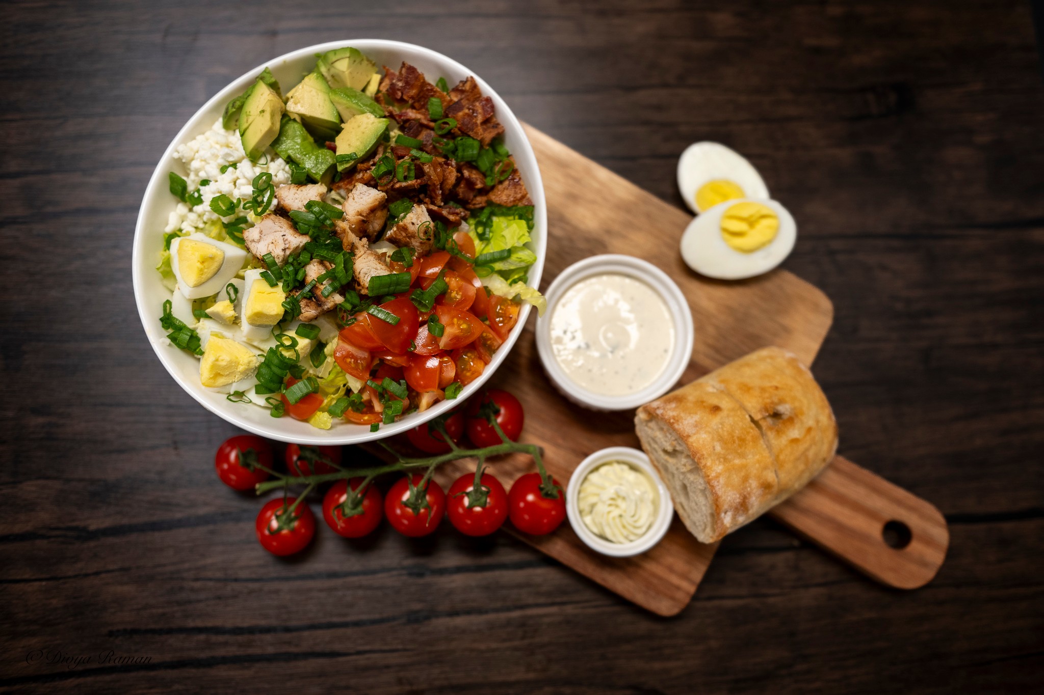 Salad bowl with avocado, tomatoes, and greens on a wooden cutting board, with cherry tomatoes, boiled eggs, and a rolled pastry nearby