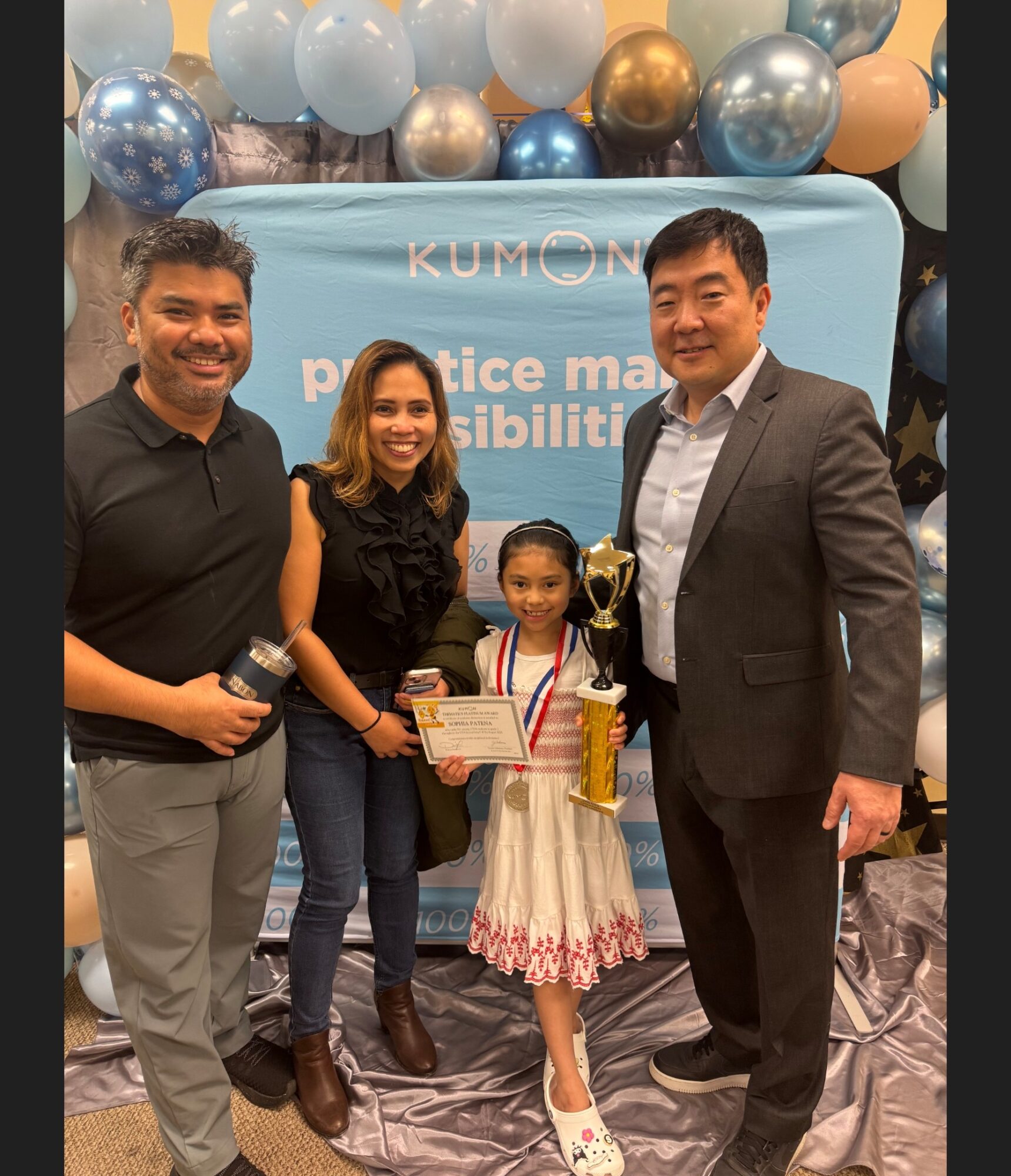 Four people, including a young girl holding a trophy and certificate, standing in front of a blue backdrop with balloons.