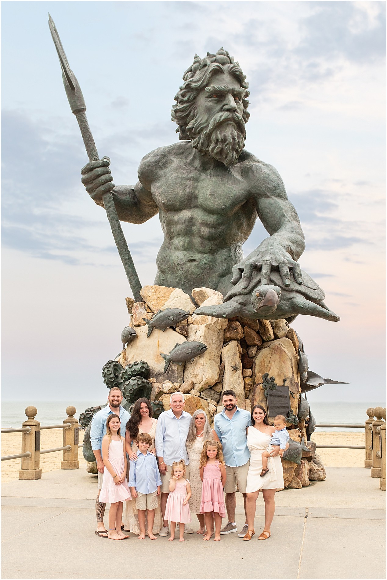 Group of people standing in front of a large statue of a bearded man holding a spear, with rocks and a carved face, on a platform with a railing.