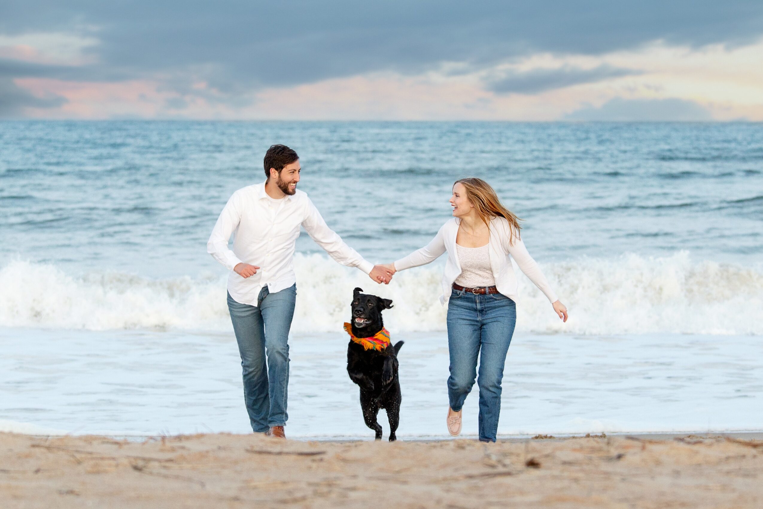 A couple holding hands with a black dog on a beach, ocean waves in the background, cloudy sky, casual clothing.