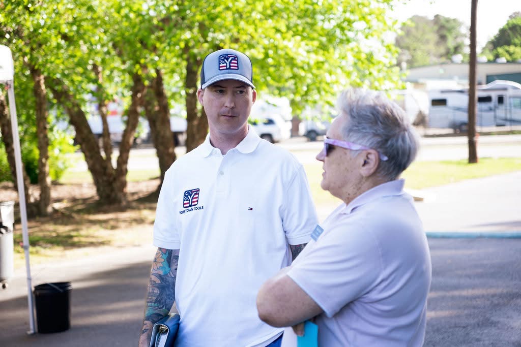 Two people talking outdoors, one young man in a white shirt and cap, and an older woman with sunglasses, trees in background.