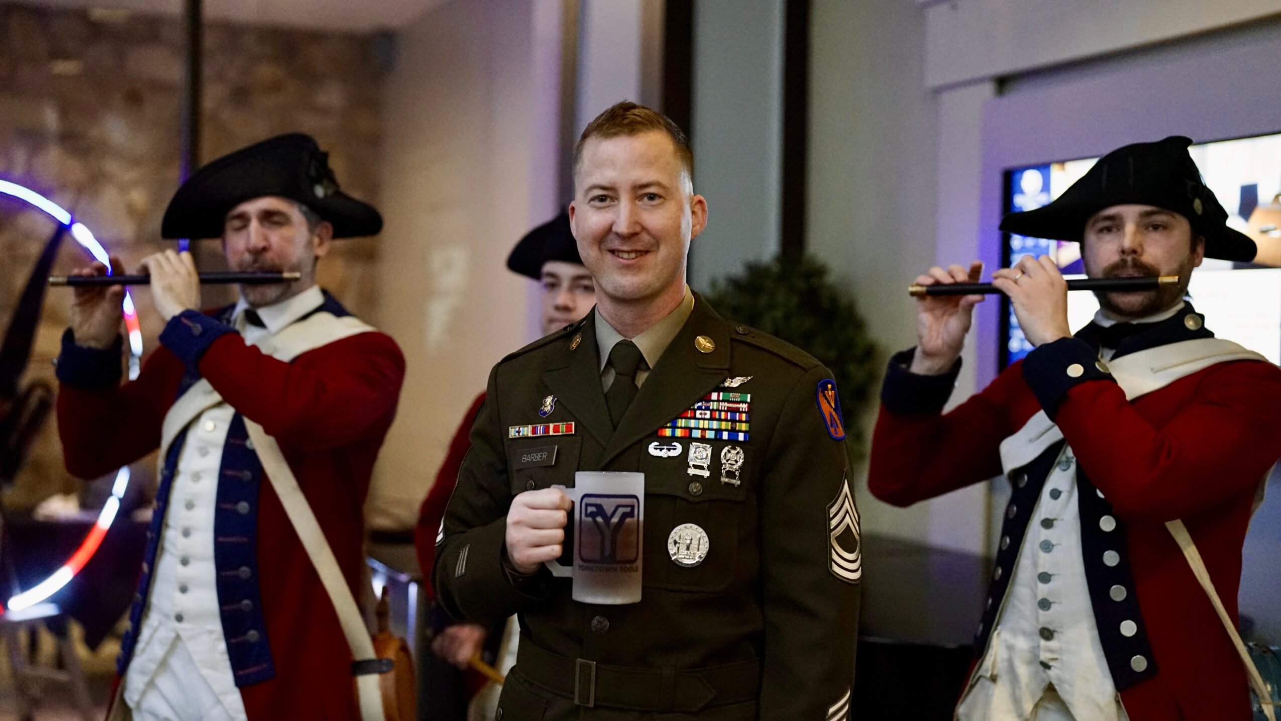 A smiling man in military uniform standing between two people dressed as historical soldiers, indoors with a modern background.
