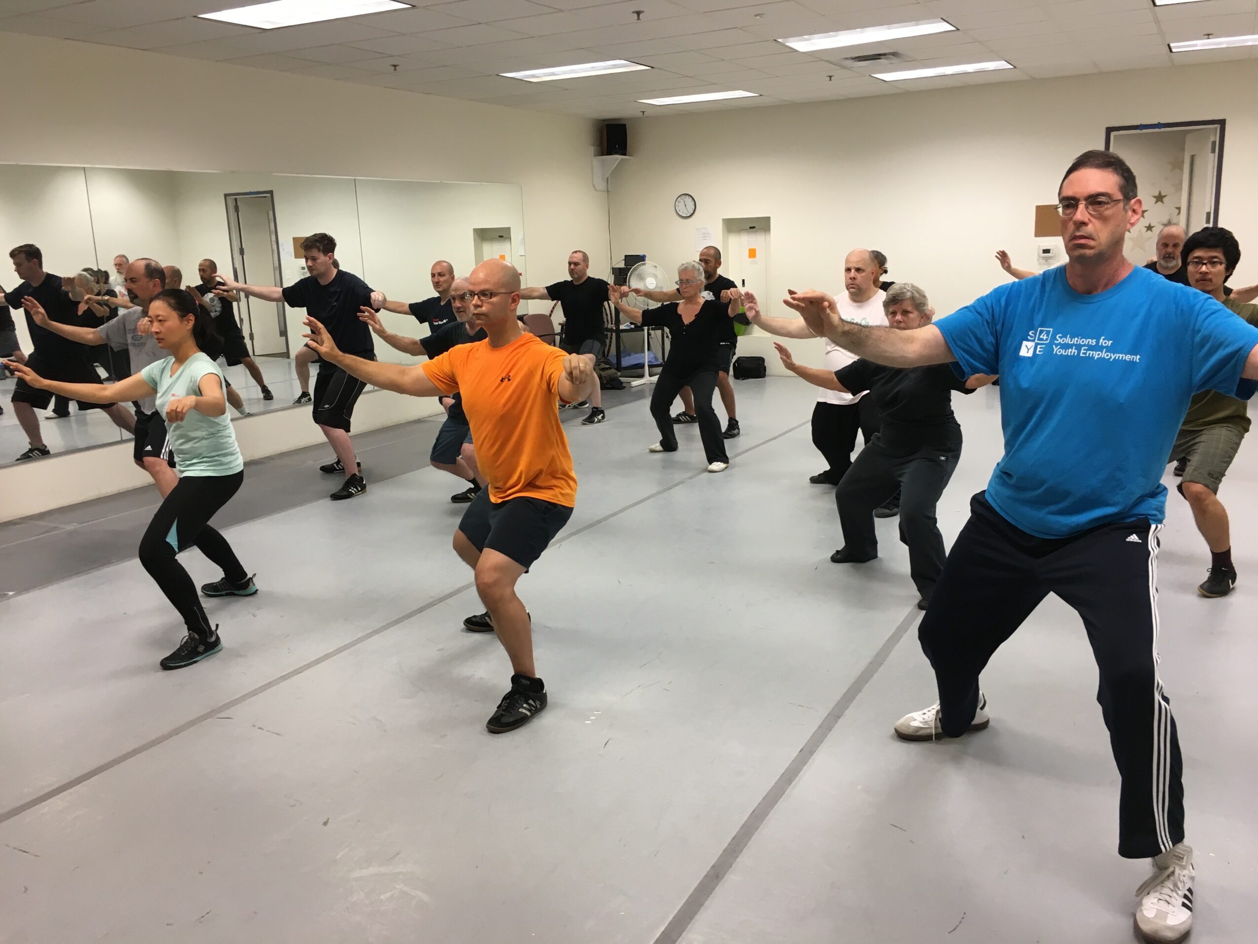 Group of people participating in a dance or exercise class in a studio with mirrors, some with arms extended, diverse ages and genders.
