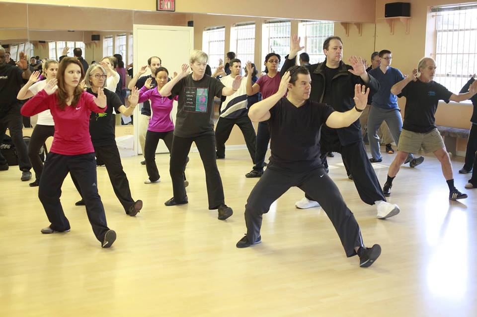 Group of people practicing dance or exercise in a studio with mirrors, some with arms raised, moving in unison.