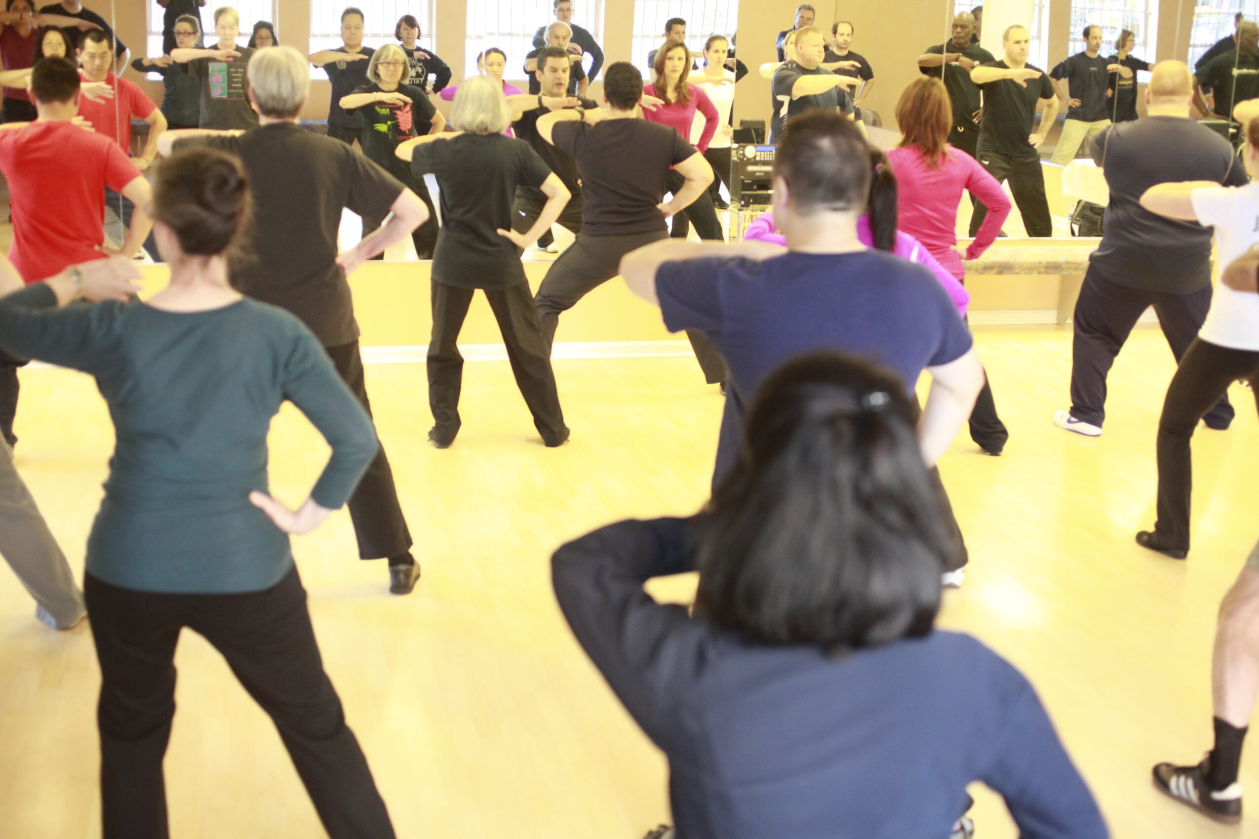 Group of people practicing dance or exercise in a studio with mirrors, facing forward, with arms raised and hands on hips.