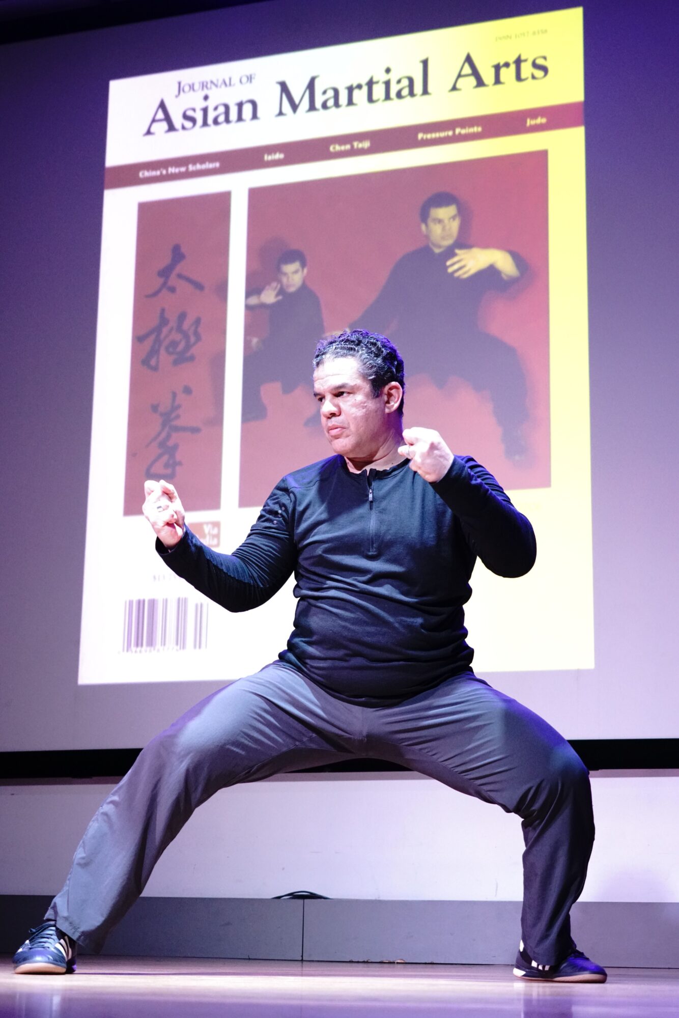 Person in black clothing performing a martial arts move in front of a display with Asian martial arts images and text.
