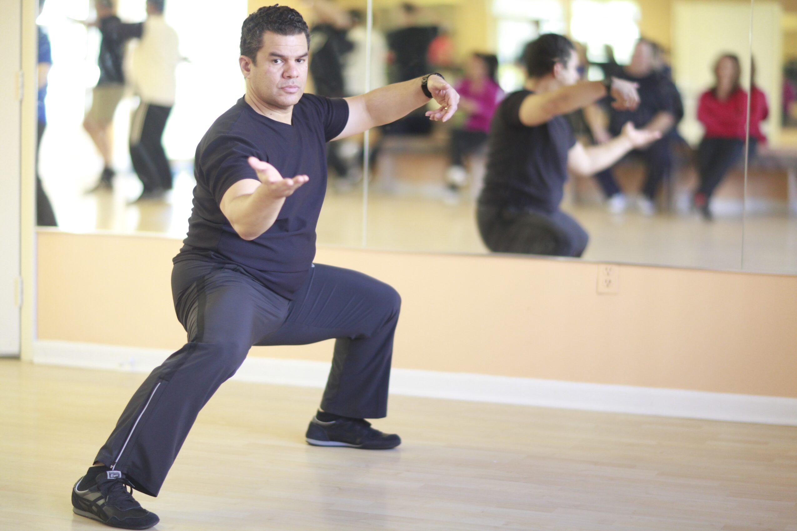 Person practicing martial arts stance in a gym with mirrors, arms extended, legs bent, focused expression.