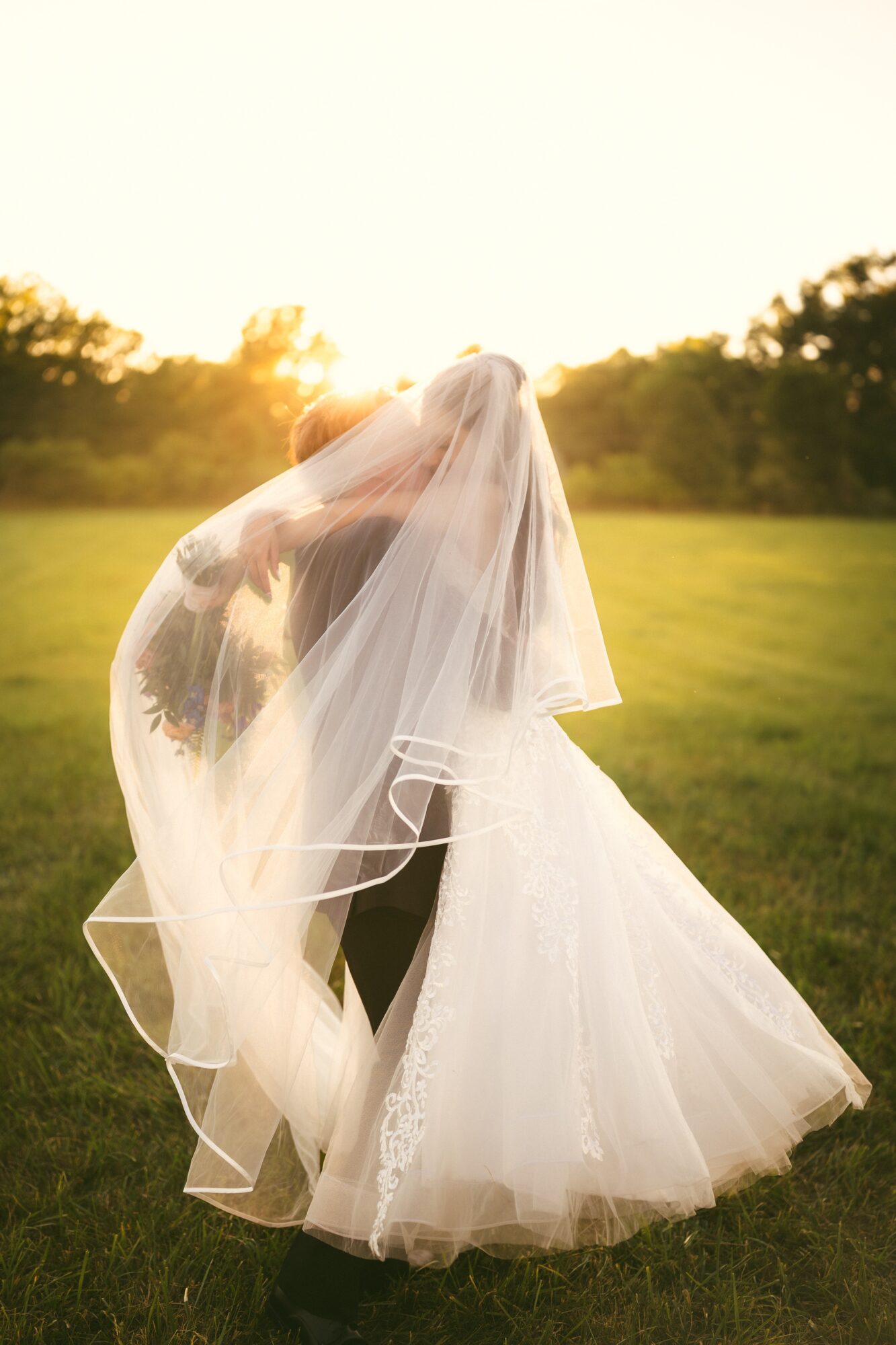 A bride and groom dancing outdoors at sunset, with the bride wearing a veil and wedding dress, in a grassy field.