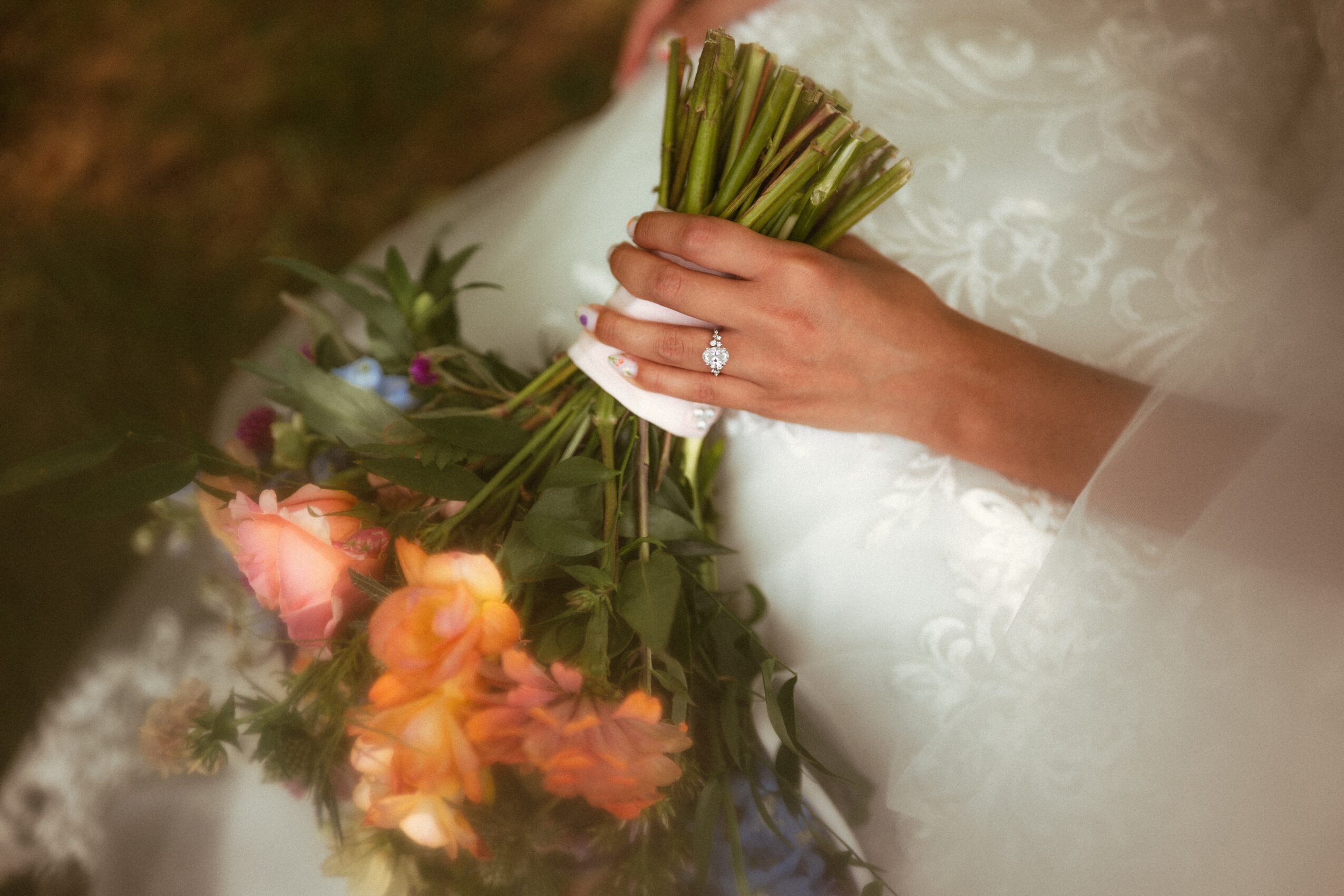 Close-up of a bride holding a bouquet of pink and orange flowers, wearing a white lace dress, with a ring on her finger.