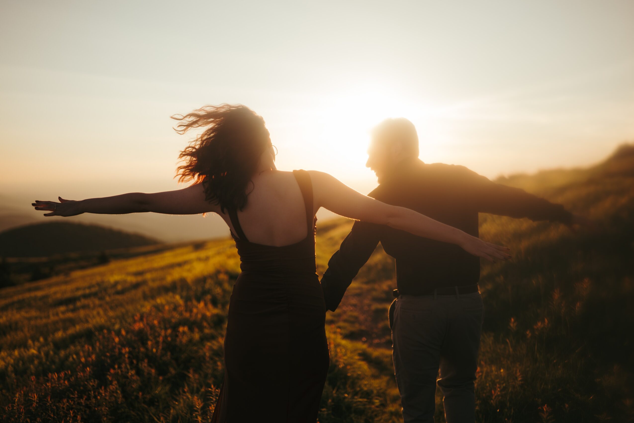 Two people holding hands outdoors during sunset, with a landscape of fields and hills in the background.
