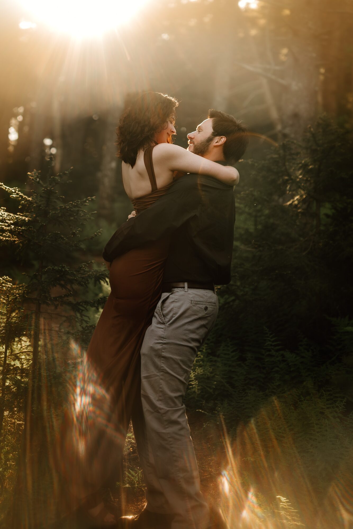 A couple embracing outdoors with sunlight filtering through trees, the woman in a dark dress and the man in a dark shirt and light pants.