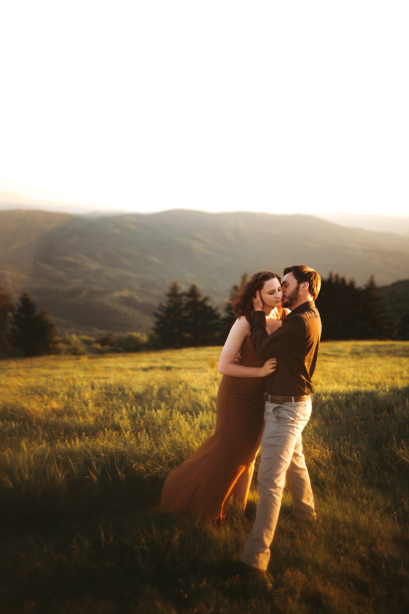 A couple stands close together in a field with mountains in the background during sunset, embracing each other.