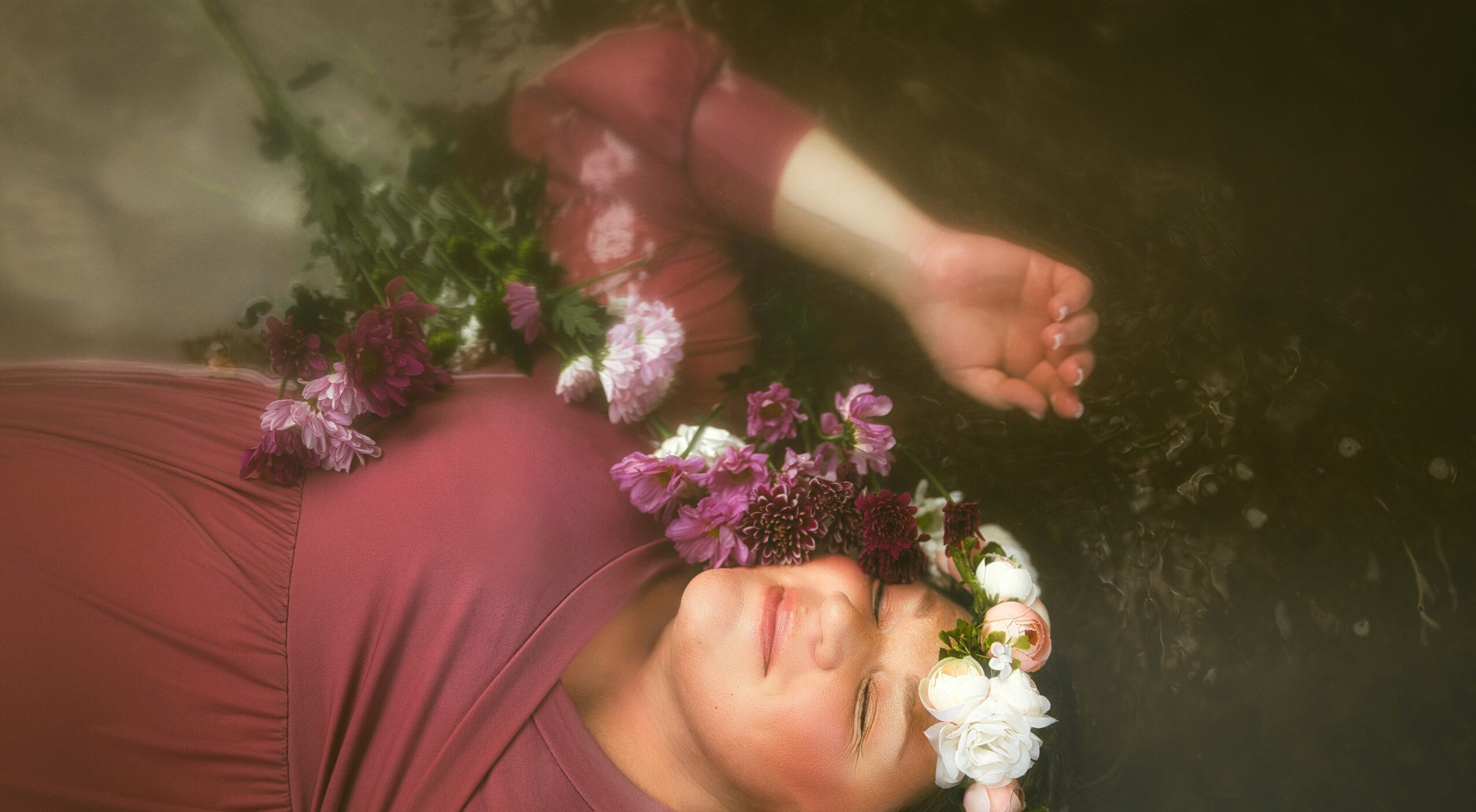 Woman with flowers on her head, lying in water with eyes closed, wearing a maroon dress.