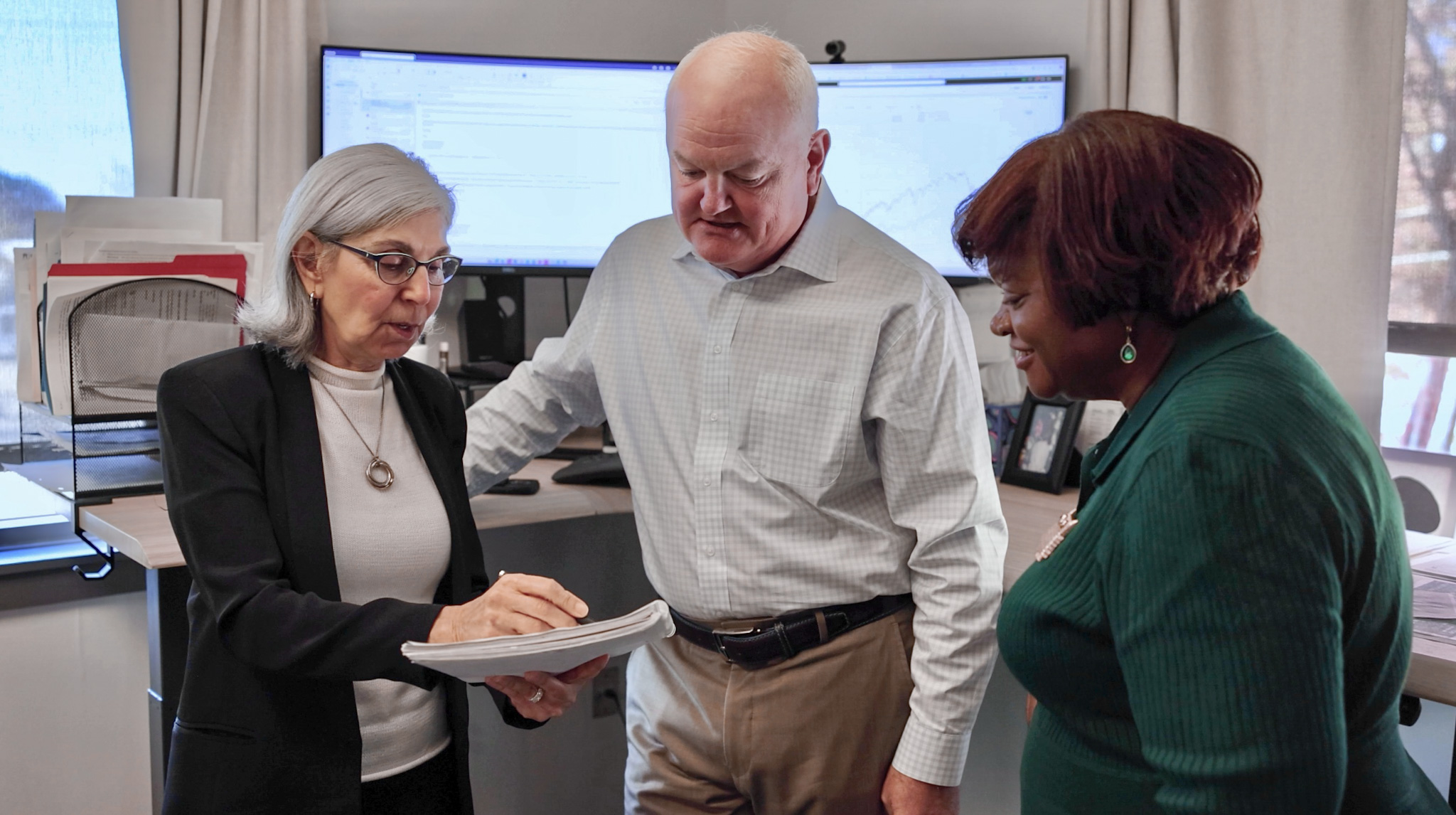 Three people in a meeting room looking at a document, with a computer monitor in the background.