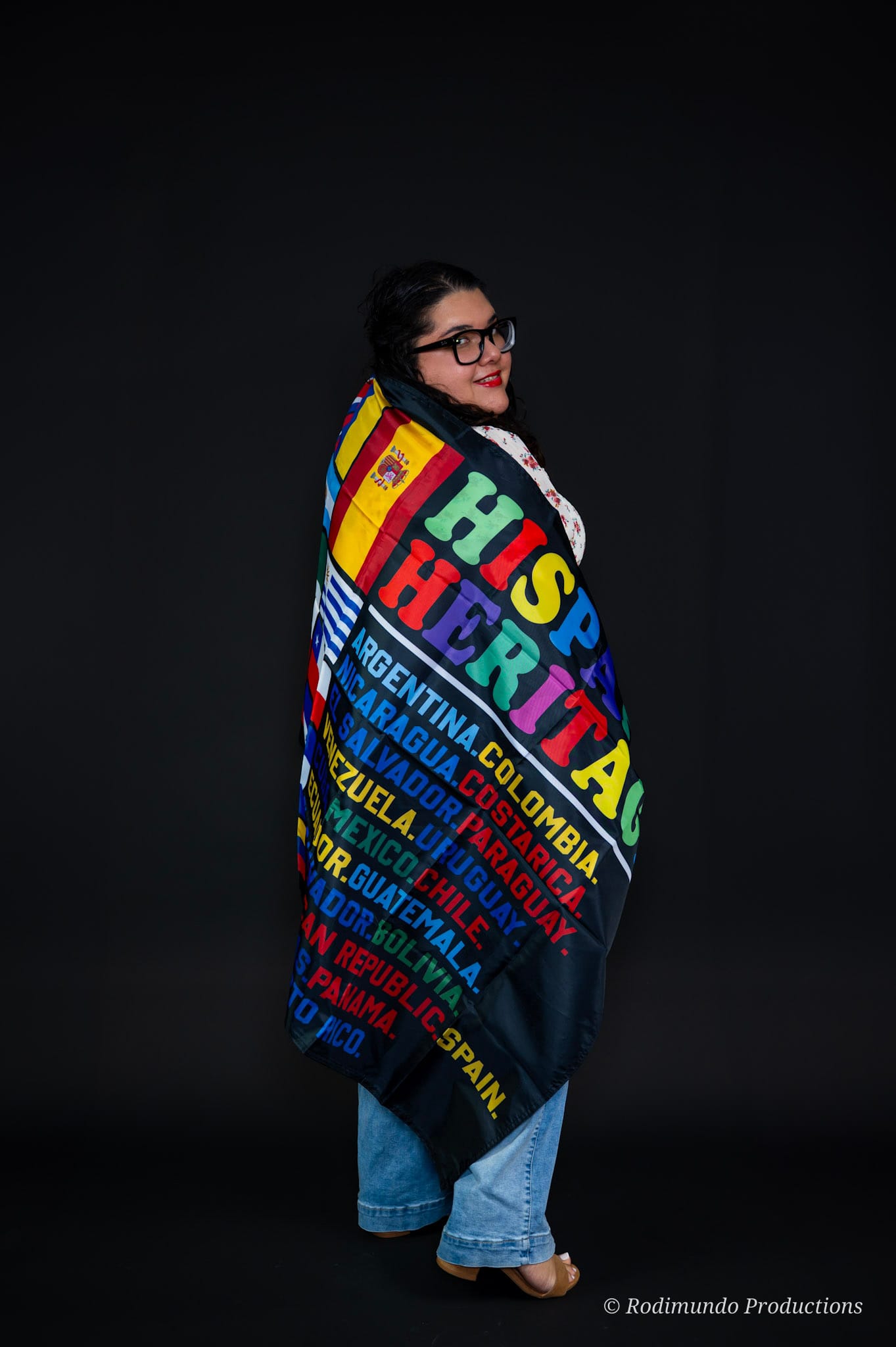 Person with glasses and dark hair wearing a colorful blanket with text, standing against a black background.