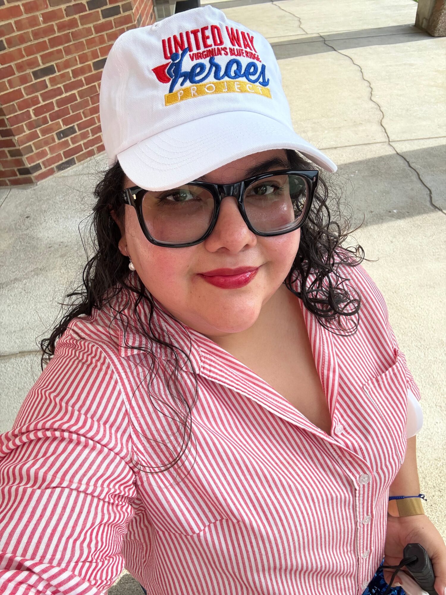 Woman wearing glasses, a white cap, and a pink striped shirt, smiling outdoors.
