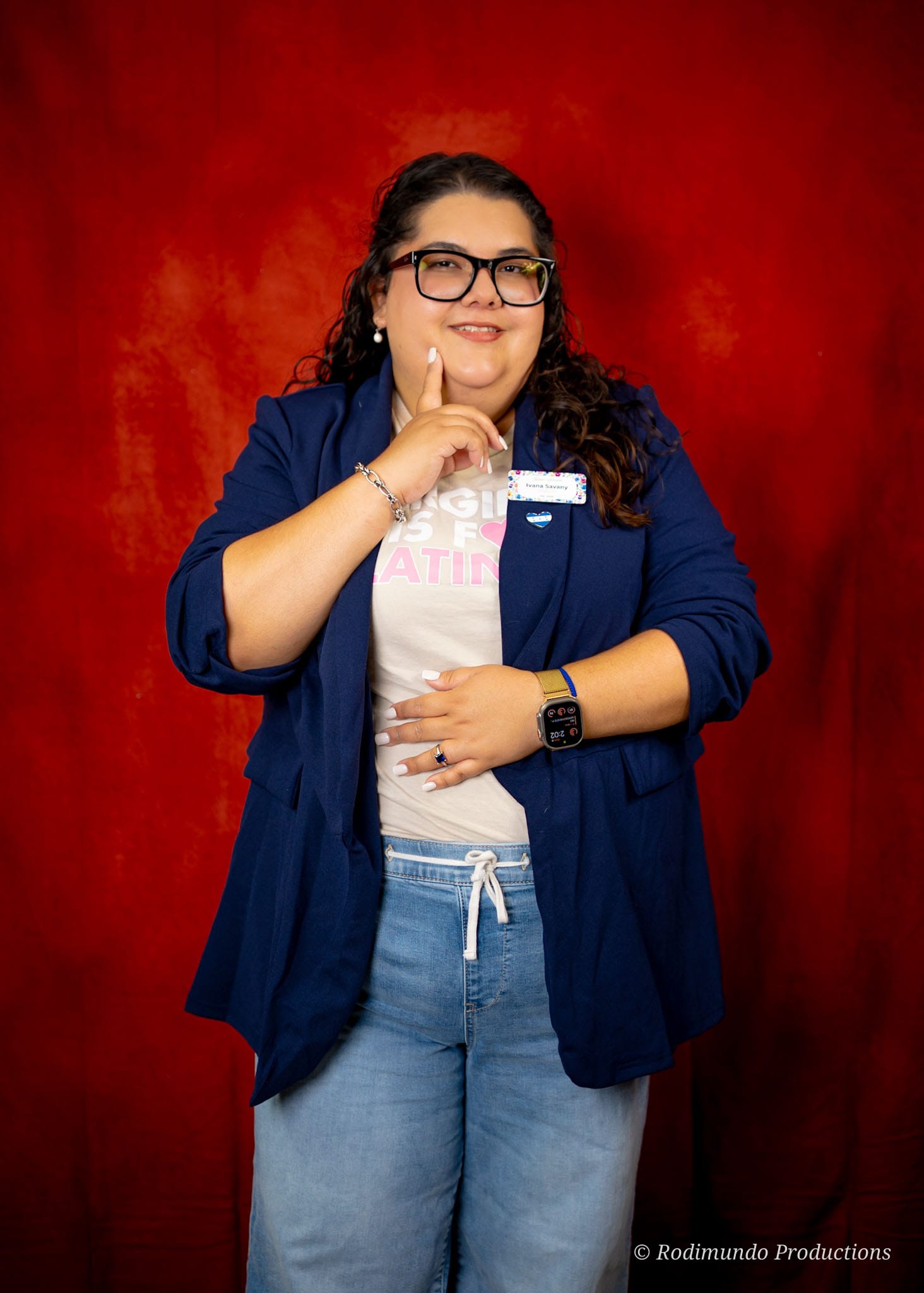 Woman with glasses, dark curly hair, wearing a blue jacket, white t-shirt, and jeans, standing against a red background.