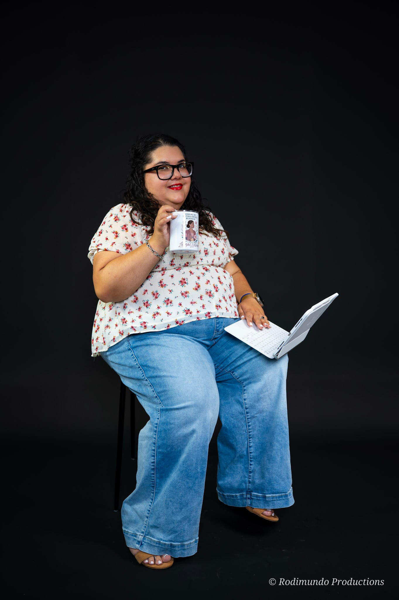 Woman with glasses, curly hair, wearing a floral top and jeans, sitting on a chair, holding a coffee mug and a laptop, against a black background.
