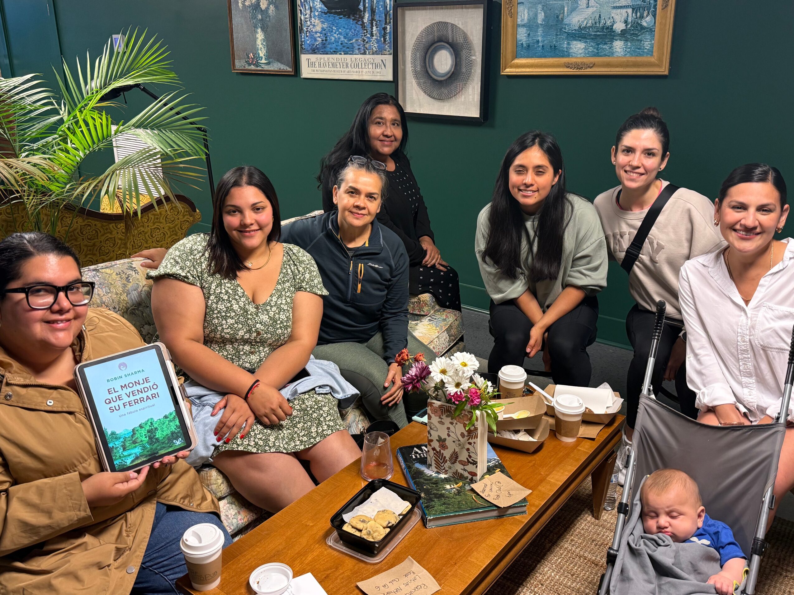 Group of seven women and a baby sitting around a table with food and drinks, smiling indoors with framed pictures on the wall.
