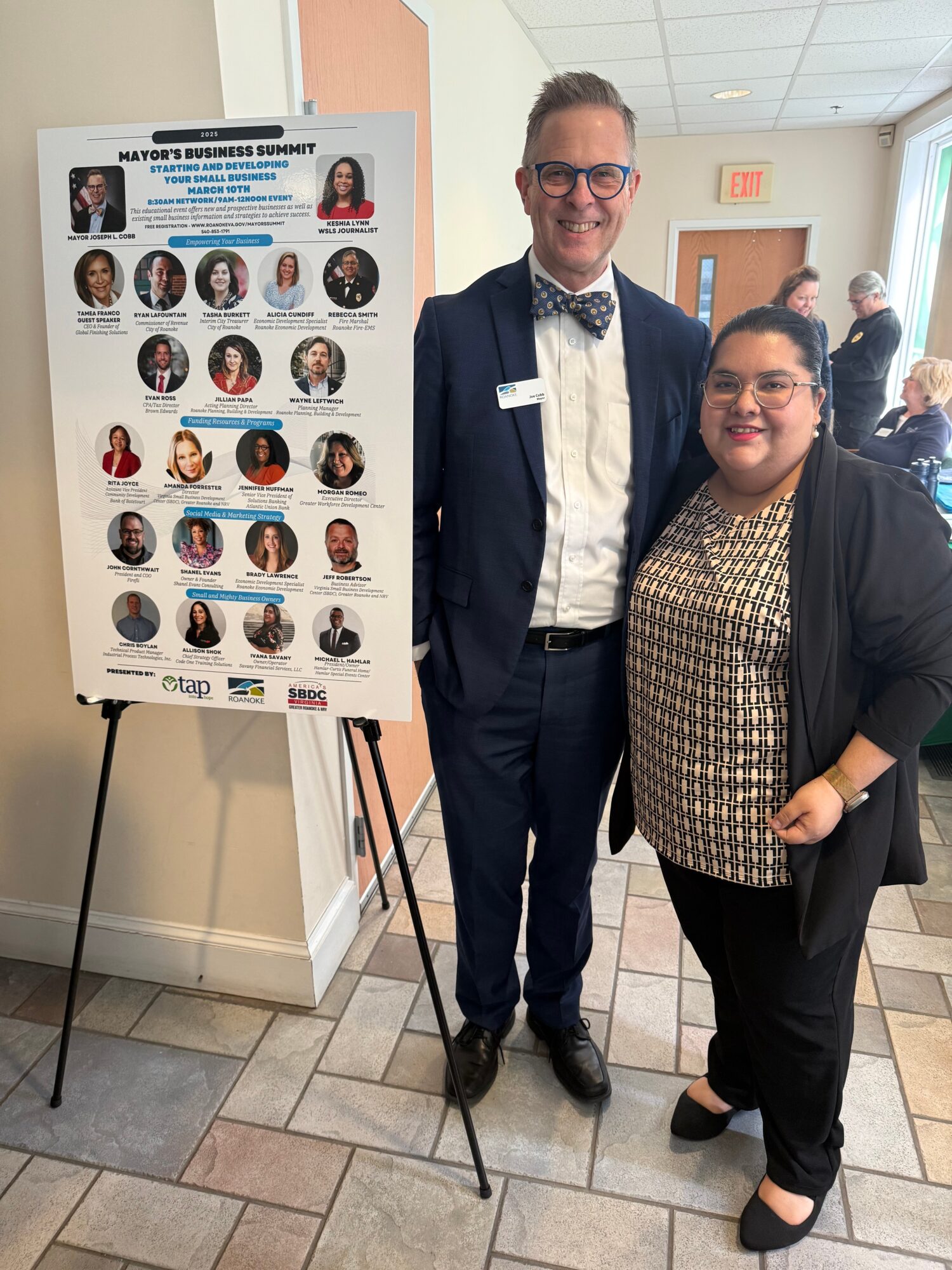 Two people standing indoors next to a poster on an easel, smiling at the camera. The man wears a suit and bow tie, the woman wears a patterned top and black jacket.