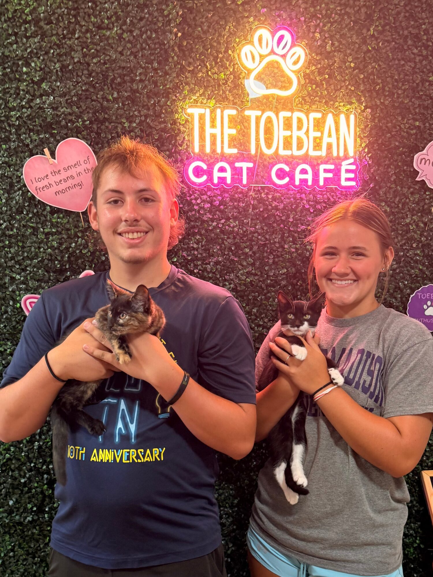 Two smiling children holding cats in front of a neon sign that reads 'The Toebean Cat Café'.