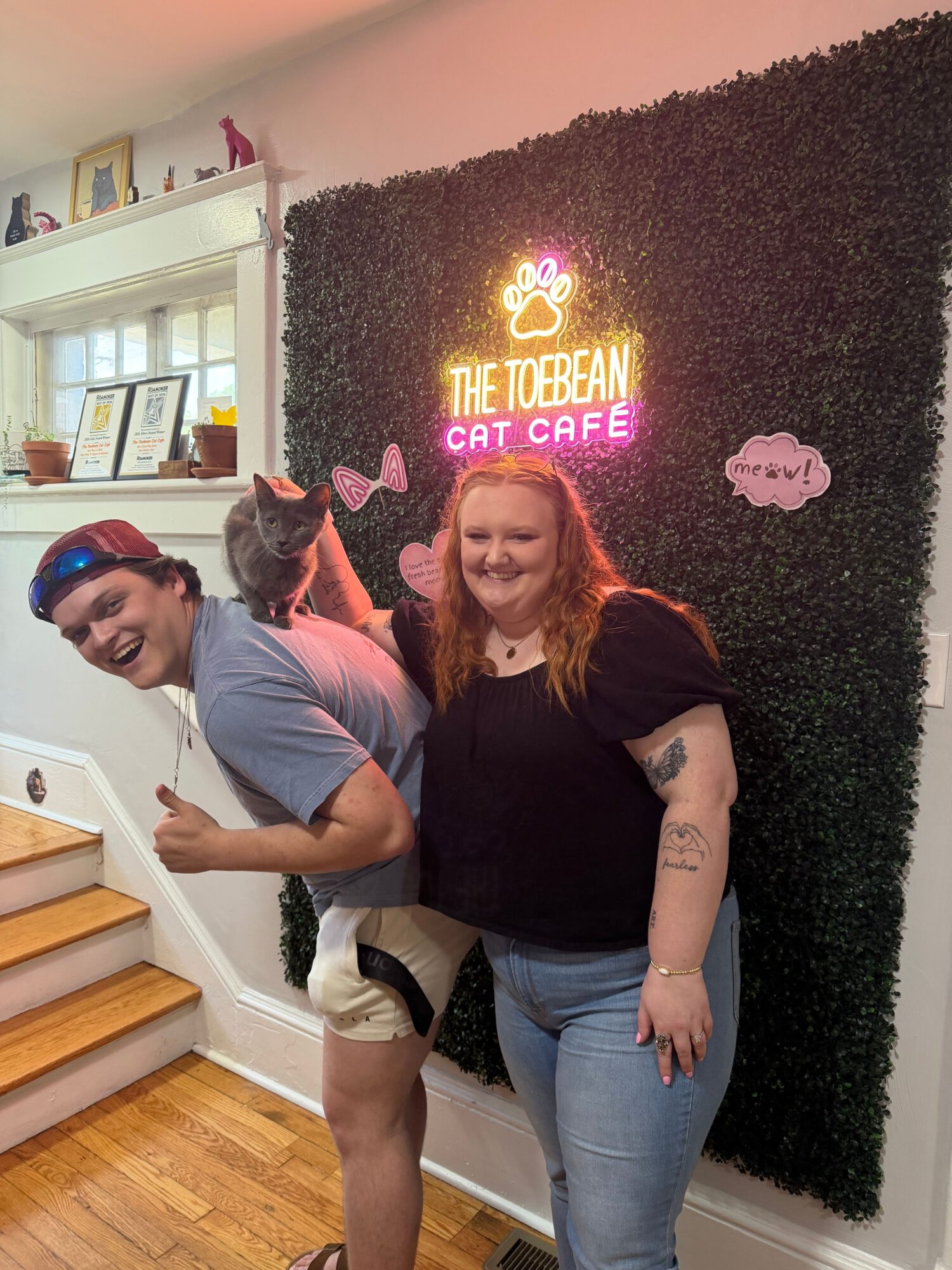 Two women and a cat in front of a black wall with neon sign, one woman with sunglasses, the other with curly hair.